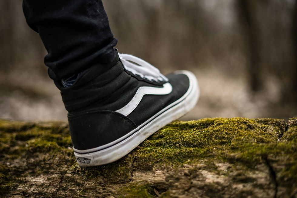 A close-up of a person wearing black high-top sneakers with a white stripe, standing on a moss-covered log in an outdoor setting. The background is blurred, featuring a natural, wooded area that creates a peaceful and earthy atmosphere.
