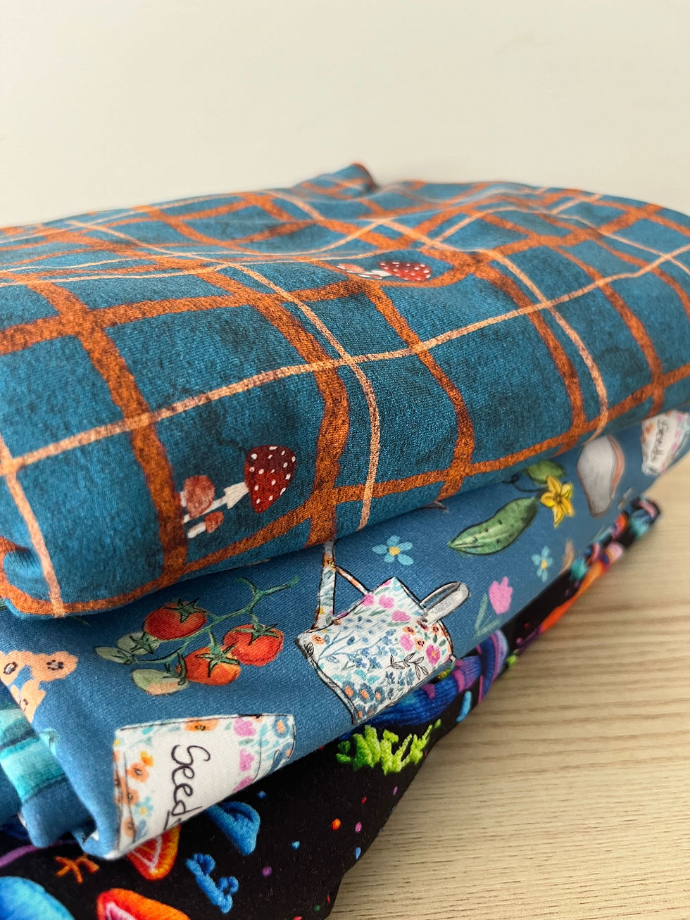 Stack of folded patterned knit fabrics on a table, with blue plaid cloth on top and a visible Seeds label; cozy, colourful textile scene