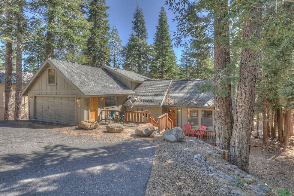 Beautiful Treehaus home in forest setting with gray roof and beige walls. Red chairs on the porch, surrounded by tall pine trees under a clear blue sky.