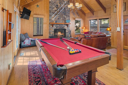 Living room with a red pool table, cues, and colorful balls. Stone fireplace, leather couches, and a wooden interior create a warm atmosphere.