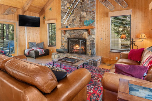 Cozy living room with a stone fireplace, crackling fire, leather sofas, wooden walls, and a vibrant red rug. Snowshoes and skis hang on walls.