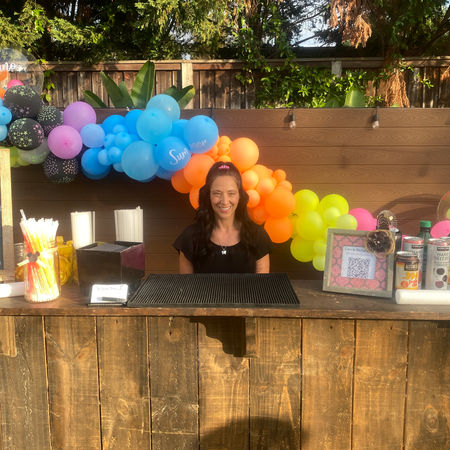 Bartender behind wooden bar with menu and bar options displayed and balloon arch behind her