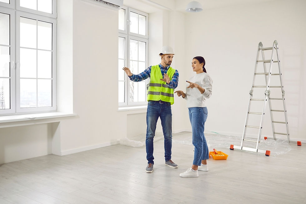 Man in hard hat and safety vest talks with woman in a bright empty room, beside a ladder and paint tray.