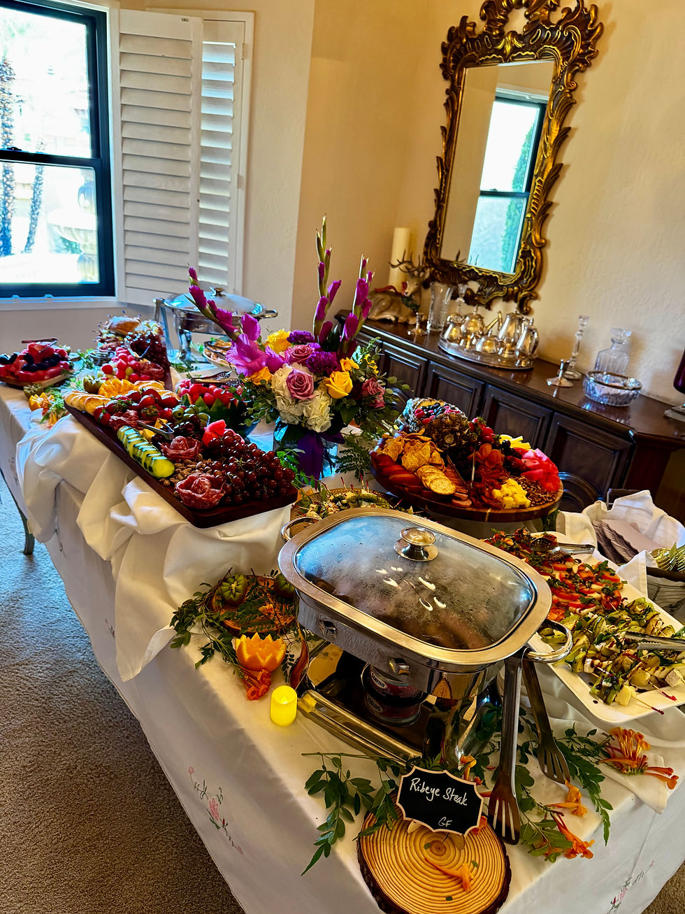 Elegant buffet table with fruit, charcuterie and hot dishes, floral centerpiece, and sign reading Ribeye Steak GF in a bright room