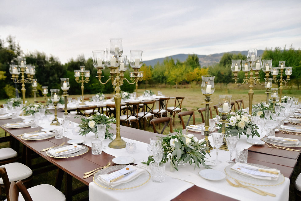 Elegant outdoor table catering setting with white linens, gold cutlery, crystal glasses, floral centerpieces, and brass candelabras, in a lush garden.
