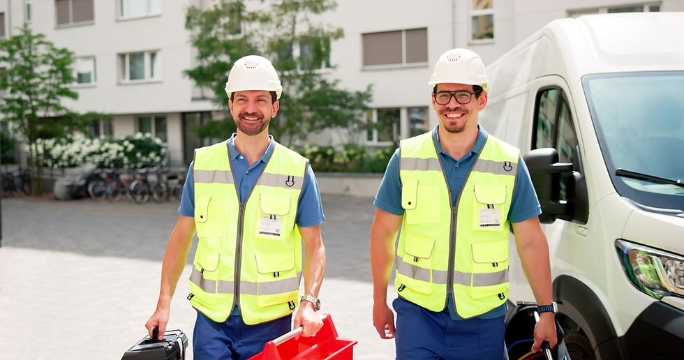 Two smiling workers in yellow vests and hard hats carry tools near a white van. Background shows a building and trees. Sunny day.