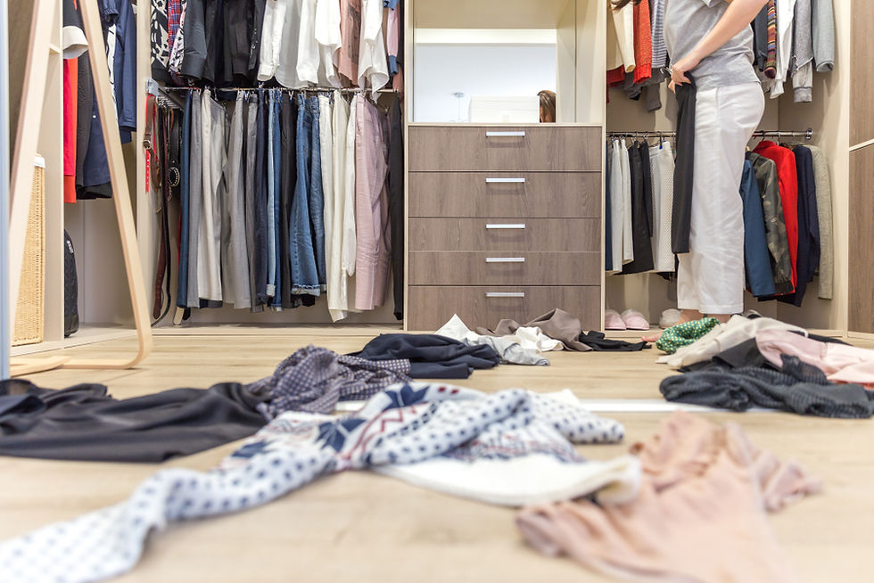 Messy closet with clothes scattered on the floor. Person holding black pants, looking through a wardrobe of various garments. Light wood floor.