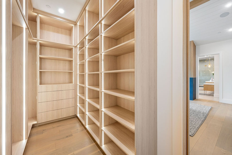 Spacious walk-in closet with empty wooden shelves and drawers, lit by soft lighting. Modern interior with a glimpse of a room beyond.