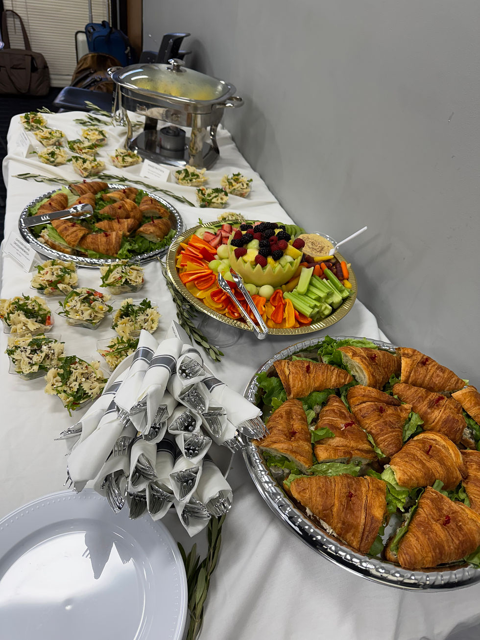 Buffet table with croissants, fruit and veggie platters, chafing dish, neatly rolled napkins, and plates in a bright room.