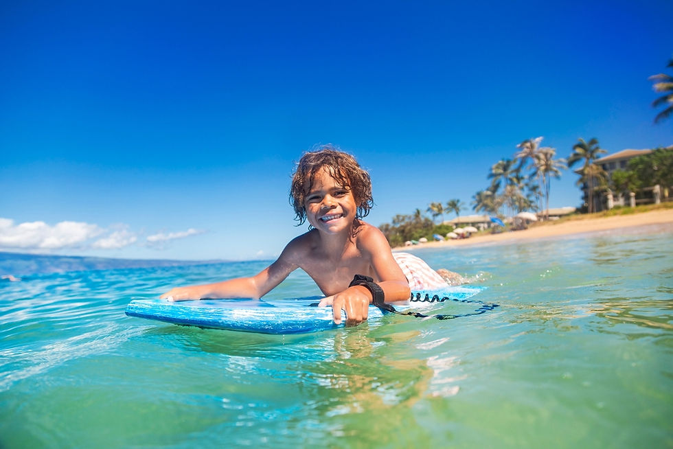 Child smiles while bodyboarding in turquoise ocean near sandy beach with palm trees under a clear blue sky. Bright, joyful scene.