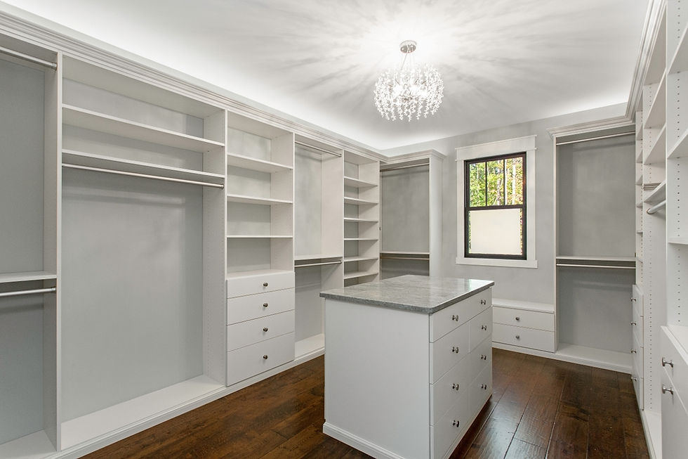 Spacious walk-in closet with white shelves and a central island, hardwood floors, and a chandelier. A window overlooks a leafy view.