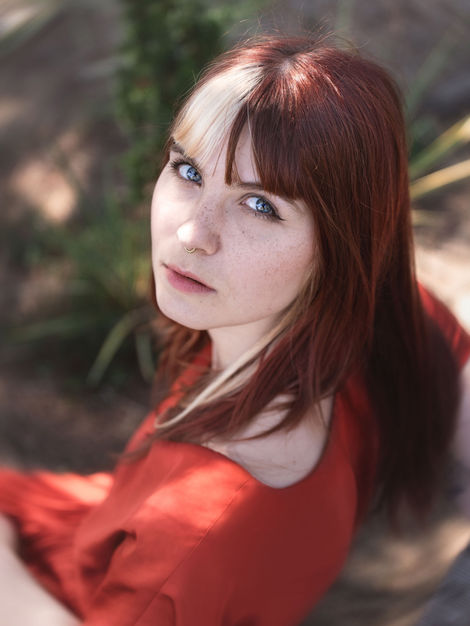 Portrait d’une femme au regard bleu intense, cheveux et robe aux tons cuivre, sur fond végétal kaki, lumière naturelle