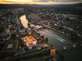 Weite Luftaufnahme der Stadt bei Sonnenuntergang mit Fluss, Brücken und dem beleuchteten Festivalgelände am Ufer.