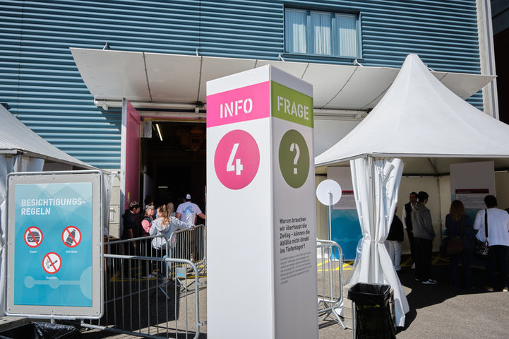 Besucher stehen vor einem Zelteingang mit Infostation und großem Info-Frage-Würfel mit der Frage: „Warum brauchen wir überhaupt die Zwilag – können die Abfälle nicht direkt ins Tiefenlager?“