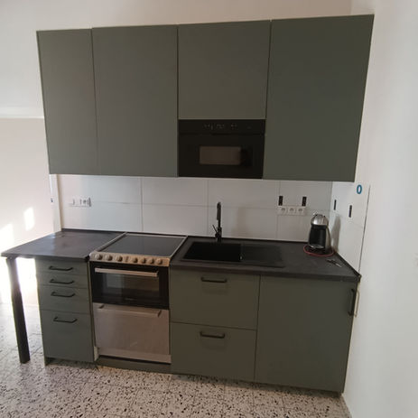 Minimalistická kuchyň s šedozelenými skříňkami, černou deskou a spotřebiči.