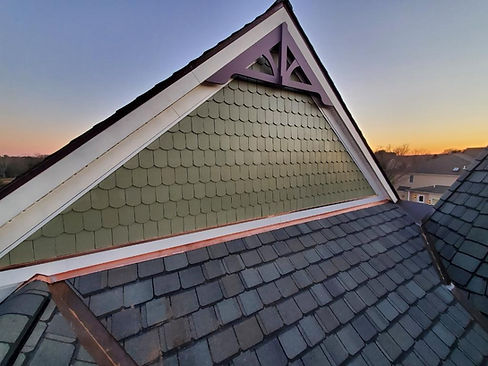 Image of a custom cedar siding home with sage green hand painted siding, copper base flashing, and grey Grand Manor shingles. The siding is smooth and even, with no visible brushstrokes. The color is a deep, rich sage green that contrasts nicely with the white trim. The siding is well-maintained and looks new. The copper base flashing provides a touch of elegance and helps to protect the siding from moisture damage. The grey luxury shingles are a classic choice that complement the siding perfectly. The overall effect is a beautiful and stylish home that is sure to turn heads.