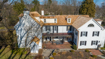 Aerial View of a Cedar Shingle Roof on a Beautiful 1740's Colonial Home, Additionally it includes White Stucco Dormers, Three Copper Flat Seam Roofs, And One Standing Seam Roof. Aerial view of a cedar shingle roof on a beautiful 1740s colonial home. The home has white stucco dormers, three copper flat seam roofs, and one standing seam roof. The roof is in good condition and shows the natural beauty of the cedar shingles. The home is surrounded by trees and shrubbery, giving it a rustic feel.