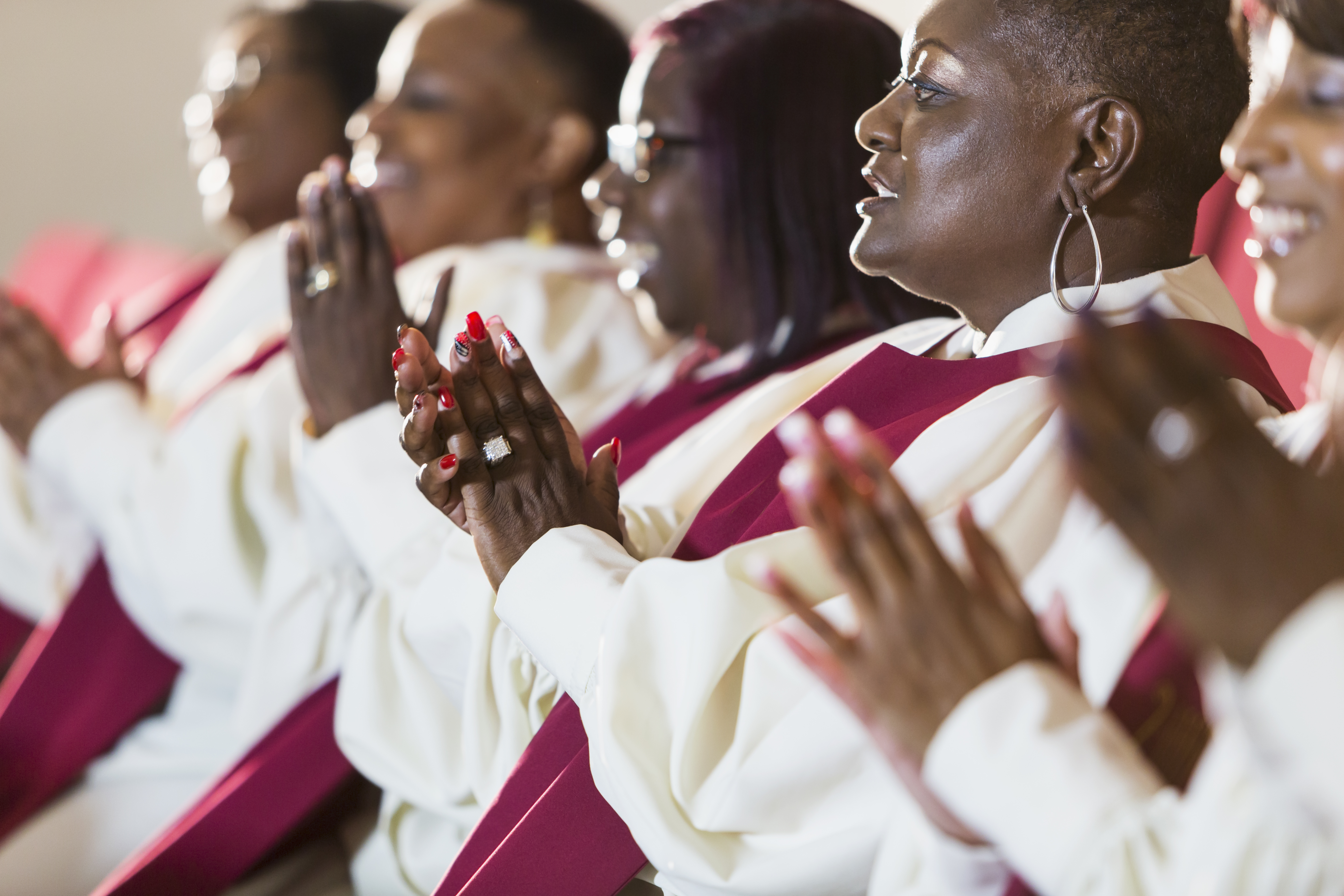 A gospel choir featuring five black women in white robes with red stoles singing and clapping their hands