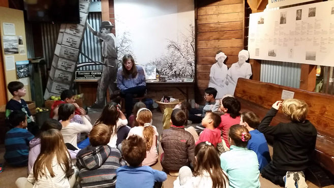 color image of children on field trip, sitting on floor while an adult speaks with them