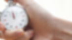 Hand holding a silver stopwatch with red and black numbers, against a light blurred background. The timer shows 15 seconds.