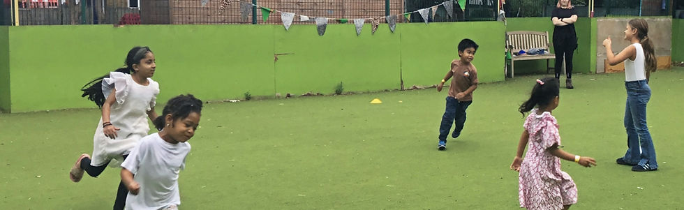 Children playing games on green field during a sunny afternoon, with a woman standing.