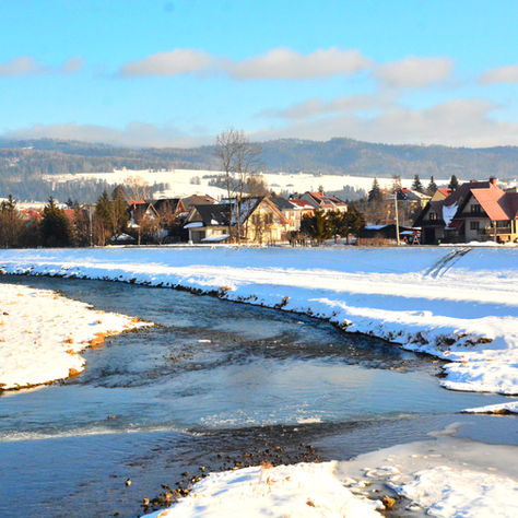 Snowy village with houses and red roofs by a winding river. Mountains in the background under a clear blue sky. Peaceful winter scene.