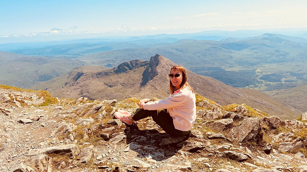 Rachel Lawrence know as The Girl with the Pilates Mat sitting on rocky mountain summit, smiling at camera under clear blue sky. Wide view of distant peaks and valleys in background.