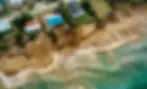 Aerial view of severe coastal erosion affecting a New Zealand beach. The image shows several houses precariously perched at the edge of a steep, eroded cliff. The beach below is partially collapsed, with sand and sediment visibly washed into the turquoise ocean waves. The erosion has dramatically altered the landscape, with sections of the coastline crumbling away, endangering the structures above.