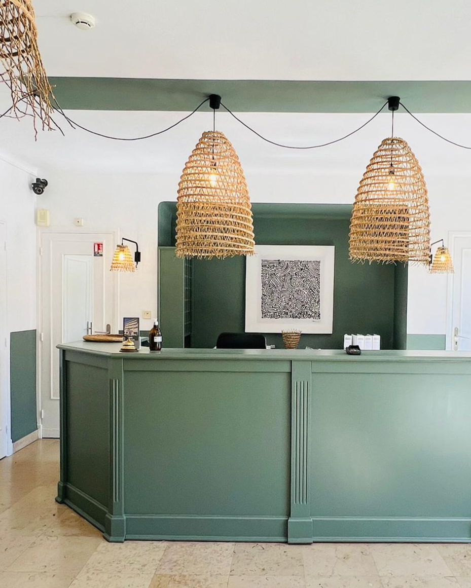 Comptoir d’accueil vert avec suspensions en rotin tressé, murs verts, sol en pierre naturelle et décoration minimaliste.