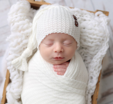 a newborn baby boy wrapped in white with a white sleepy hat posed in a crate with an all white set