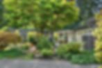 Midcentury modern house designed by architect Roland Terry with large windows, surrounded by lush greenery, trees, and a garden path. Bright, tranquil outdoor setting.