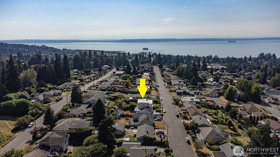 Aerial view of a waterfront suburban neighborhood with a yellow arrow pointing to a house, under a calm blue bay sky.