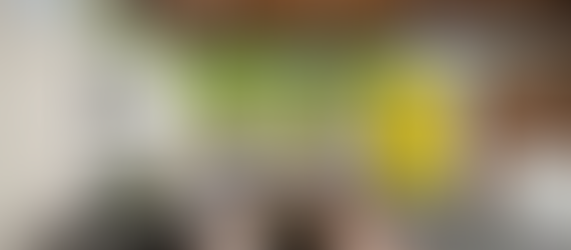 Midcentury modern living room with a gray sectional sofa, colorful cushions, abstract art, and a plant. Large windows show trees and a yellow door. Architect Mary L. Davis