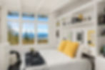Bright bedroom with large window view of trees and mountains. Bed with yellow pillows, black blanket, bookshelves, and framed art. Serene mood.