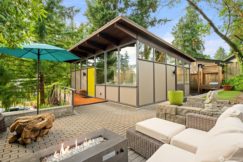 Modern patio with a fire pit, white seating, and a wooden sculpture. Surrounded by trees, a house with yellow door and large windows. Relaxing vibe. Architect Mary L. Davis