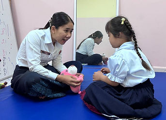 Image of Cambodia female teacher sitting on the floor with a girl both looking at each other interacting. A mirror shows a reflection behind and the teacher is holding a pink stuffed animal.