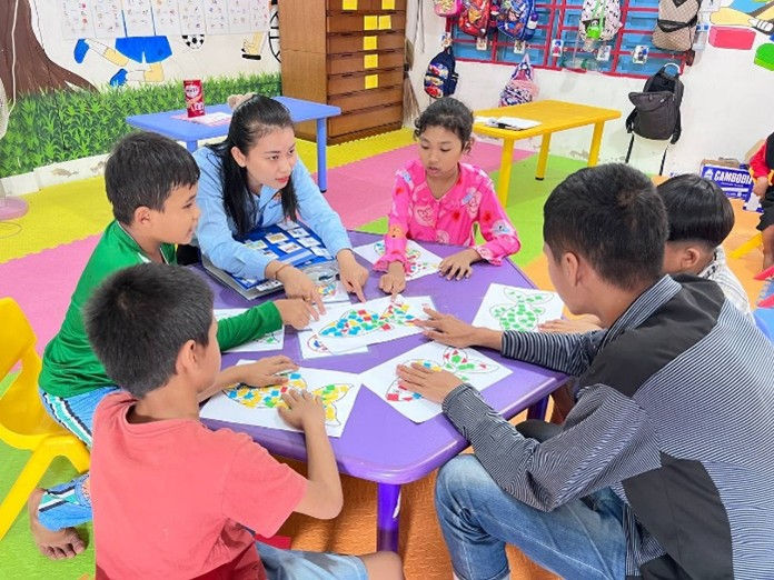 Daneth at the table with students seated in a circle all looking at her talking and pointing to colorful art in the middle