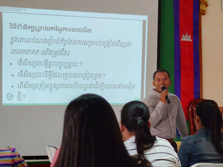 Cambodians man smiles and speaks into microphone in front of Khmer banner and slide with Khmer script