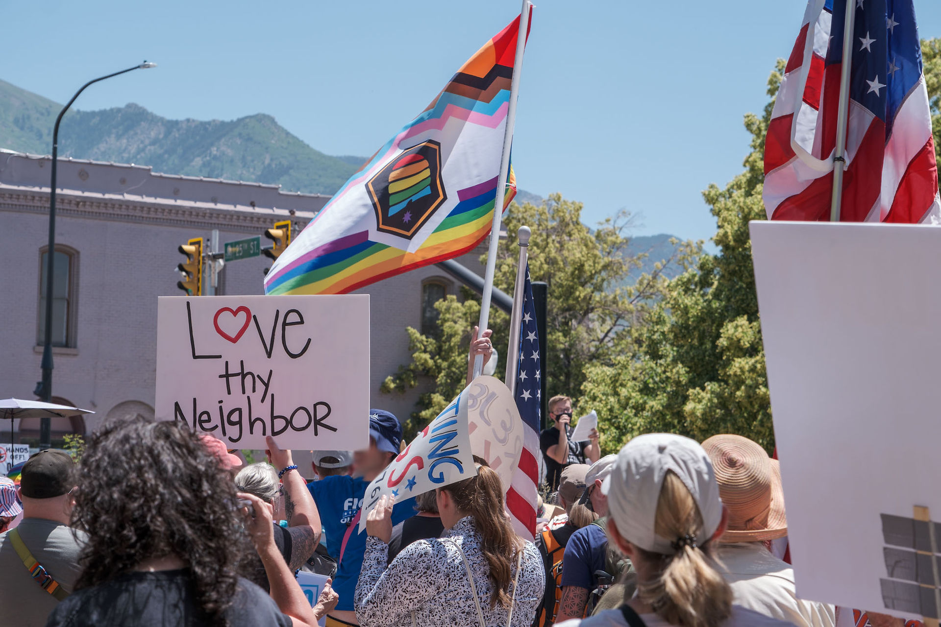 A sign reads “Love thy neighbor.” Beside it, a Utah flag in Pride colors flies. Several U.S. flags are also visible.