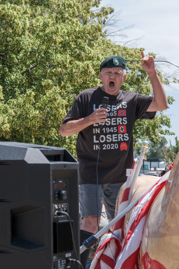 A man in a military beret speaks into a microphone while making large gestures.