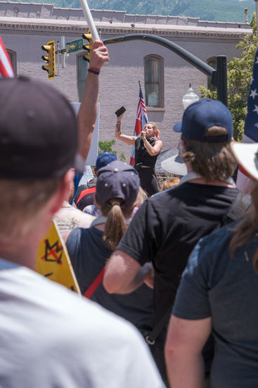 A young woman in black speaks into a handheld microphone while making large gestures with her other hand, which holds a cell phone.