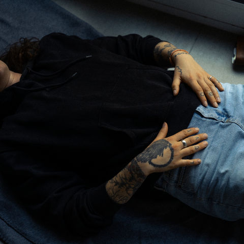 Person liegt im Innenraum am Boden, von Hals bis Oberschenkel fotografiert, trägt Jeans und schwarzen Pulli, Hände liegen flach auf dem Unterbauch mit sichtbaren Tattoos – Teil einer Kailo Nature Therapy Sitzung.