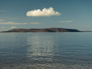 A lone swimmer in calm water beneath a single cloud, with a low island on the horizon.