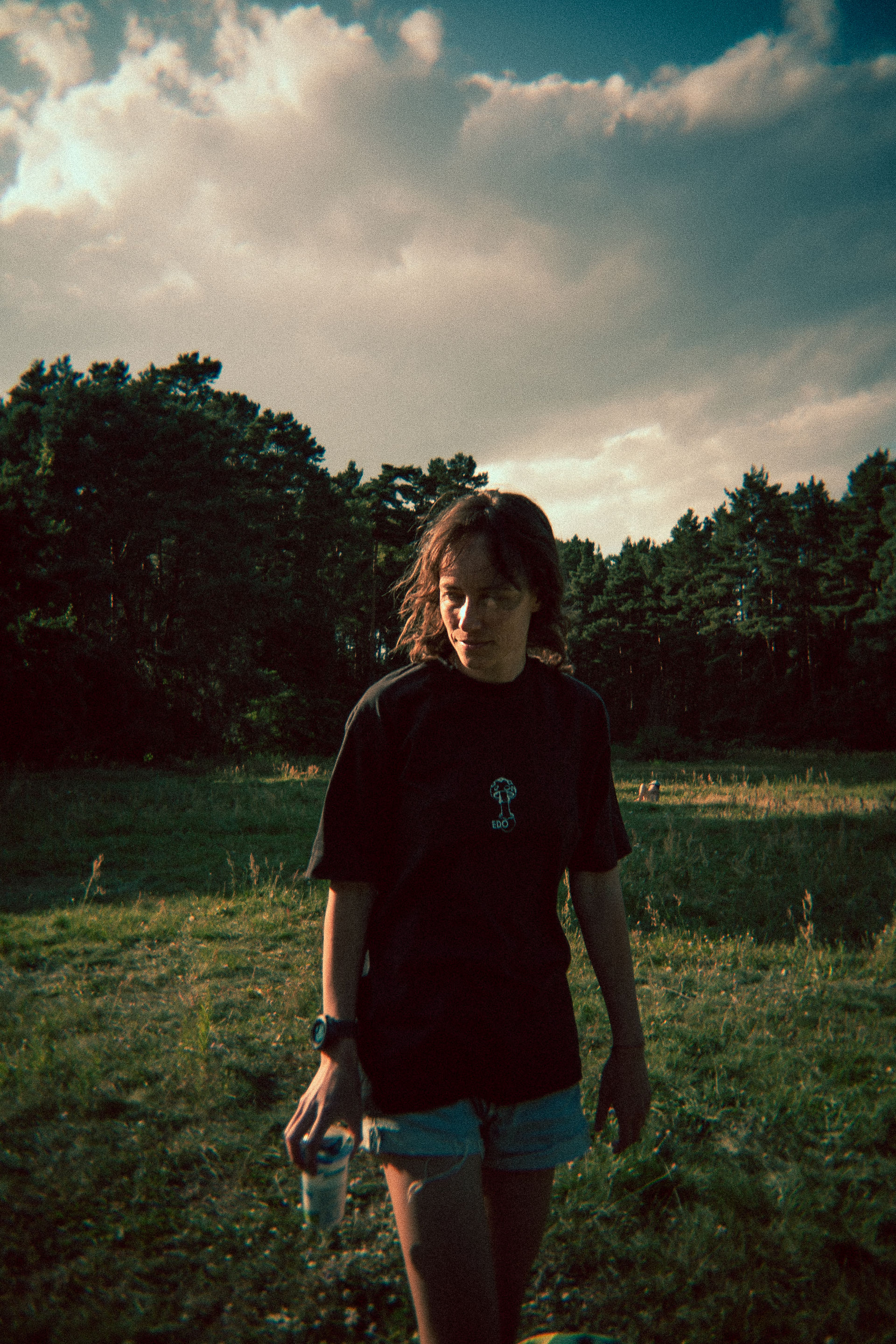 Woman in black T-shirt and denim shorts walks through a grassy field at sunset, holding a drink, under dramatic cloudy sky.