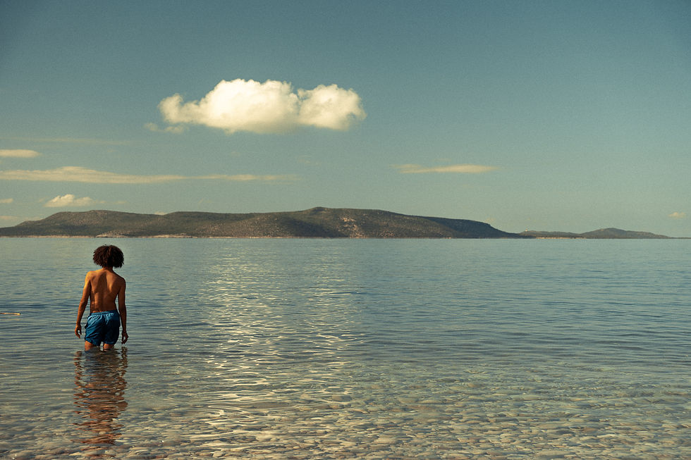 Back view of a person wading in calm clear sea, with distant low islands and a lone cloud under a pale sky.