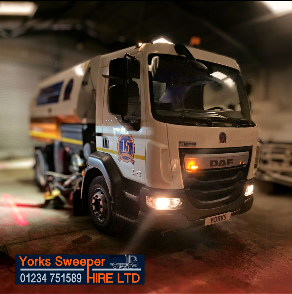 A road sweeper truck with headlights on is parked indoors. The truck is white with blue and orange details. Text: "Yorks Sweeper Hire Ltd 01234 751589". Safety Lights