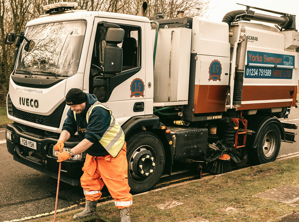 Worker in orange gear and safety vest using tools beside a street sweeper truck. Overcast day with trees in background. Truck marked "Yorks Sweeper."