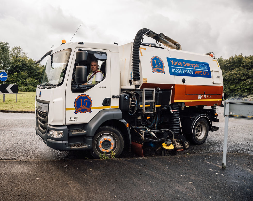 Street sweeper truck on road with a person inside, surrounded by greenery. The truck has signage with "Yorks Sweeper Hire" and a phone number.