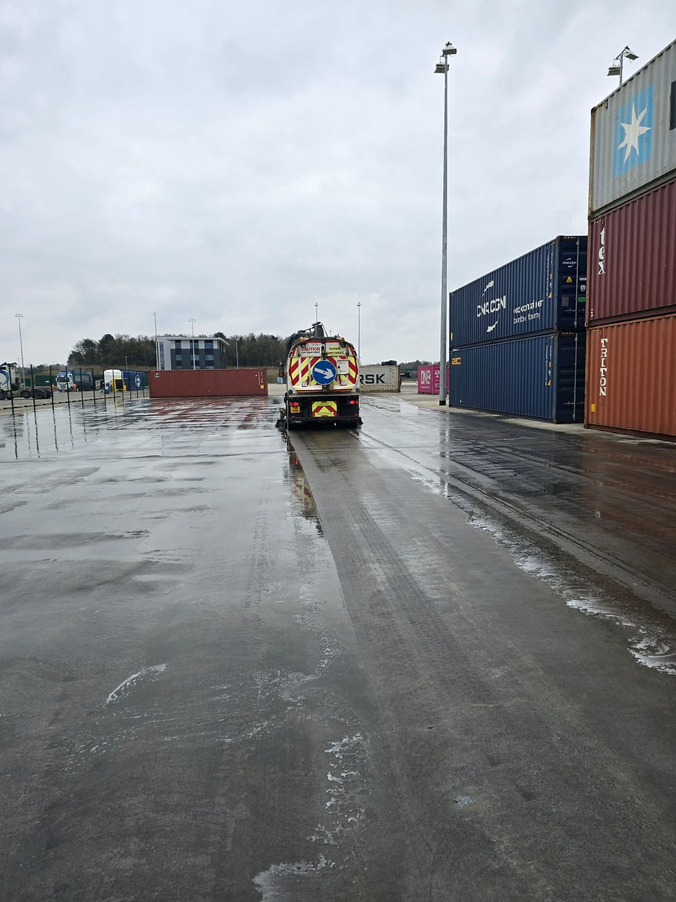 Road Sweeper on wet dockyard road, surrounded by stacked shipping containers under a cloudy sky. Arrow signs on truck. Industrial, overcast mood.