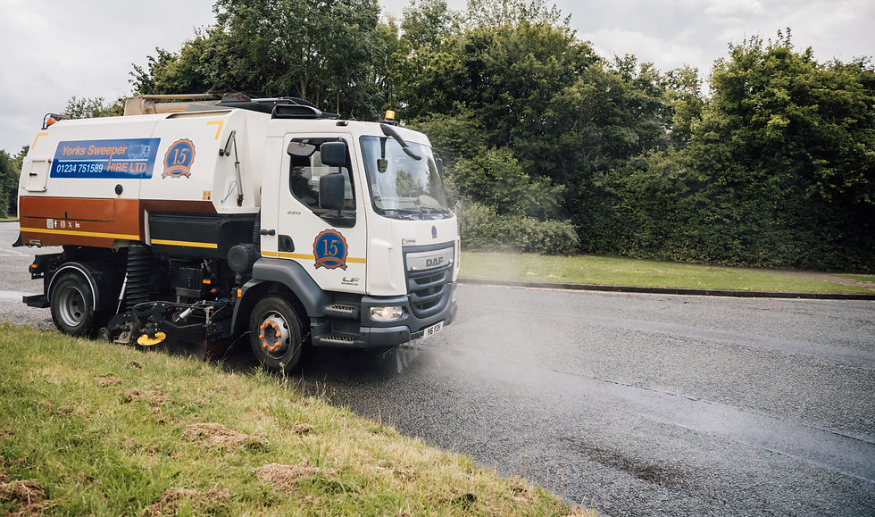 Street sweeper cleaning a road, spraying water. White truck with "Yorks Sweeper Hire Ltd" text. Green trees in the background.